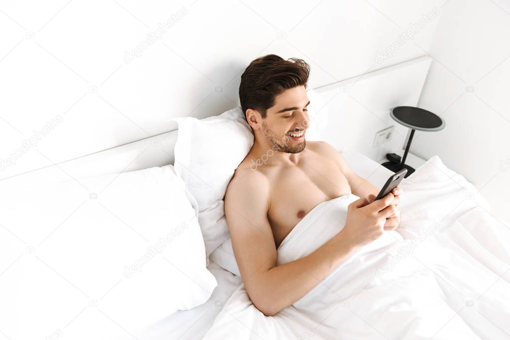 Smiling shirtless man using mobile phone while laying in bed at home