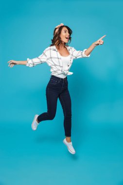 Tam uzunlukta portresi atlama bir mutlu genç ev hanımı, işaret uzakta mavi arka plan üzerinde izole