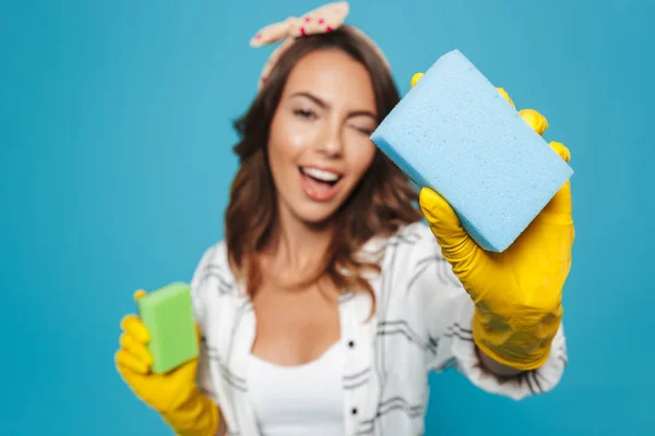 Portrait closeup of positive housewife 20s in yellow rubber gloves for hands protection holding two sponges during cleaning isolated over blue background