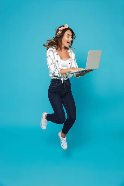 Full length portrait of a cheerful young housewife using laptop computer while jumping isolated over blue background