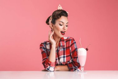 Güzel esmer pin-up kızı masada bir milkshake ile pembe arka plan üzerinde oturan ekose gömlekli portresi