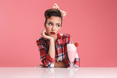 Sıkılmış bir esmer pin-up kızı masada bir milkshake ile pembe arka plan üzerinde oturan ekose gömlekli portresi