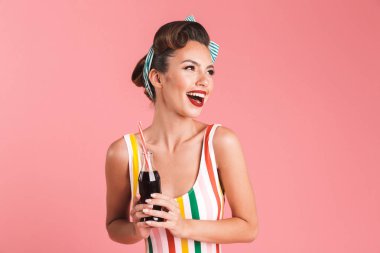Soda elinde pembe bir arka plan duvar üzerinde izole heyecanlı genç pin-up kadın görüntüsü. Kenara gülüyor arıyorsunuz.