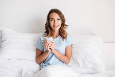 Sabahları evde yatakta otururken kupa tutan gülümseyen genç kadın