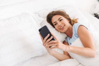 Gülümseyen genç kadın yatakta döşeme sırasında cep telefonuyla