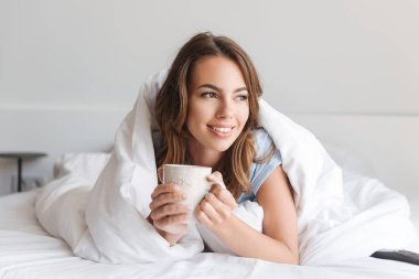 Sabahları evde yatakta döşeme sırasında kupa tutan gülümseyen genç kadın
