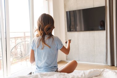 TV uzaktan kumanda ve tv izlerken kahve fincanı tutan yatağa oturan genç kadın arkadan görünüm