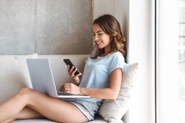 Dizüstü bilgisayar ile evde bir kanepede otururken cep telefonuyla gülümseyen genç kadın