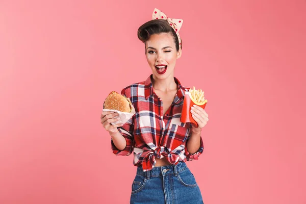 一个开朗美丽的黑发女人的肖像在头带吃薯条和一个汉堡孤立的粉红色背景 — 图库照片