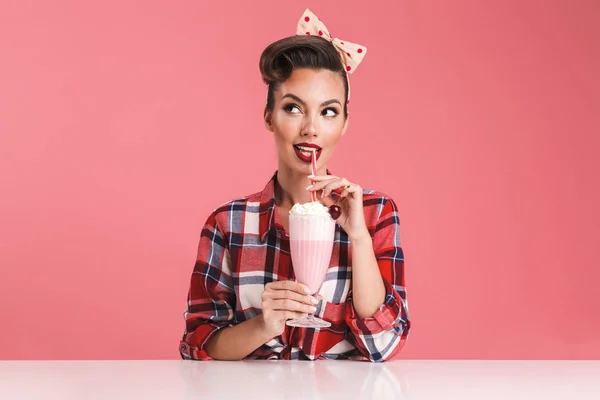 ミルクセーキを押しながら テーブルに座っているとピンク背景によそ見格子縞のシャツの笑顔ブルネット ピンナップ ガールの肖像画 — ストック写真