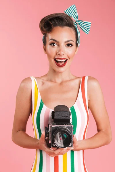 Portrait of a smiling brunette pin-up girl in plaid shirt holding vintage photo camera over pink background