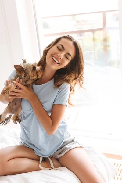 Smiling young woman holding dog while sitting on bed at home
