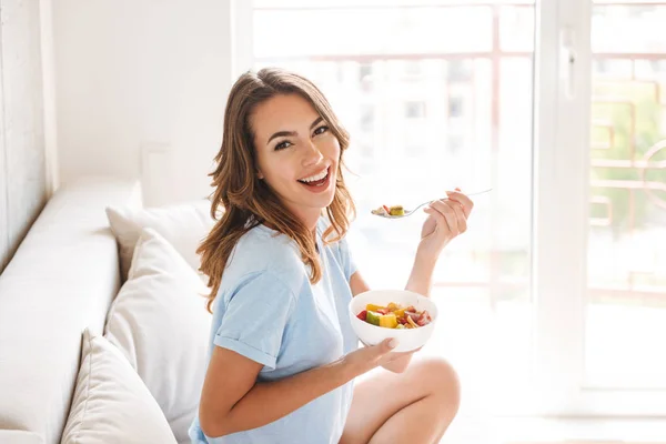 快乐的年轻妇女吃健康的早餐 而坐在沙发上在家 — 图库照片