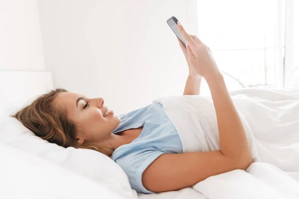 ベッドに敷設しながら携帯電話を使用して幸せな若い女 — ストック写真