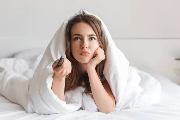 Bored young woman laying in bed covered in blanket holding tv remote control