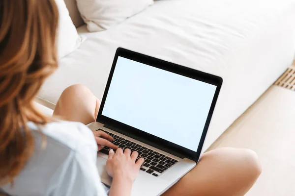 Back view of a young woman typing on blank screen laptop computer while sitting on a couch at home
