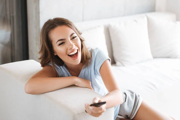 Close up of a laughing young woman relaxing on a couch at home and holding tv remote control