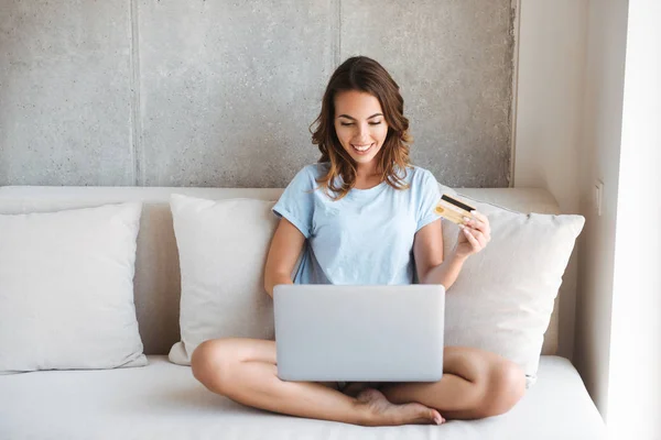 Happy young woman holding credit card while sitting on a couch at home with laptop computer