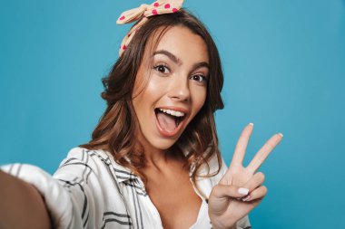 Gülümseyen ve barış işareti mavi arka plan üzerinde izole selfie çekerken gösterilen komik güzel kadın 20'li yaşlarda giyen kafa bandı portresi