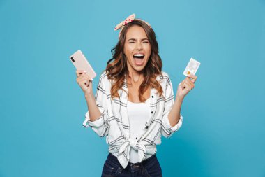Smartphone ve kredi kartı üzerinde mavi arka plan izole tutarken heyecanlı sevimli kız 20'li yaşlarda kafa bandı giyen bağırma portre