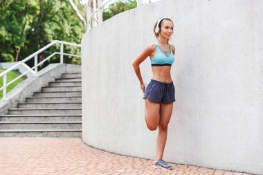 Kulaklıklar açık havada duvara germe egzersizleri yapıyor spor giyim giymiş genç sporcumuz gülümseyen