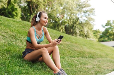 Mutlu genç sporcumuz cep telefonu açık havada çim üzerinde otururken dinlenme kulaklık spor giyim giymiş