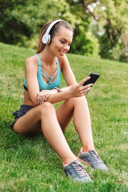 Neşeli genç sporcumuz cep telefonu açık havada çim üzerinde otururken dinlenme kulaklık spor giyim giymiş