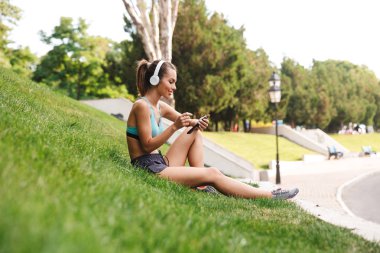 Genç sporcumuz gülümseyen cep telefonu açık havada çim üzerinde otururken dinlenme kulaklık spor giyim giyinmiş