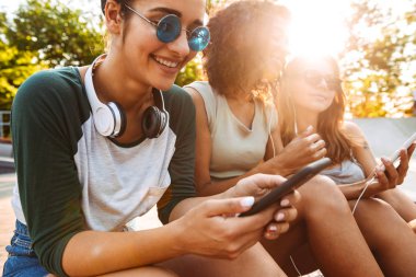 Cep telefonunu kulaklık ile müzik dinleme kullanarak birbirleriyle konuşan genç mutlu üç arkadaş kadın açık havada oturma görüntüsünü.