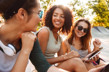 Cep telefonunu kulaklık ile müzik dinleme kullanarak birbirleriyle konuşan genç mutlu üç arkadaş kadın açık havada oturma görüntüsünü.