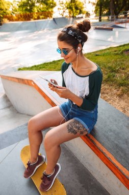 Açık havada kaykay ile müzik dinleme cep telefonuyla earphones ile oturan genç neşeli güzel kadın fotoğrafı.