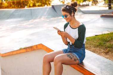 Cep telefonu kullaranarak earphones ile açık havada dinleme müzik oturan genç gülümseyen güzel kadın fotoğrafı.