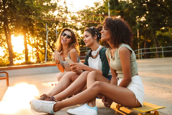 年轻快乐三朋友的形象户外与滑板坐在一起使用手机聊天 — 图库照片