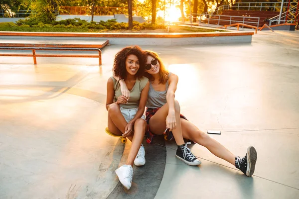 两个微笑的年轻女孩坐在公园里玩滑板的乐趣 — 图库照片