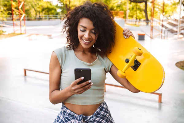 Leende Unga Afrikanska Flicka Med Mobiltelefon Stående Med Skateboard Utomhus — Stockfoto