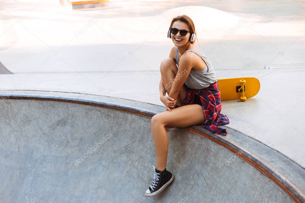 Pretty young girl in headphones listening to music while sitting with skateboard at a park