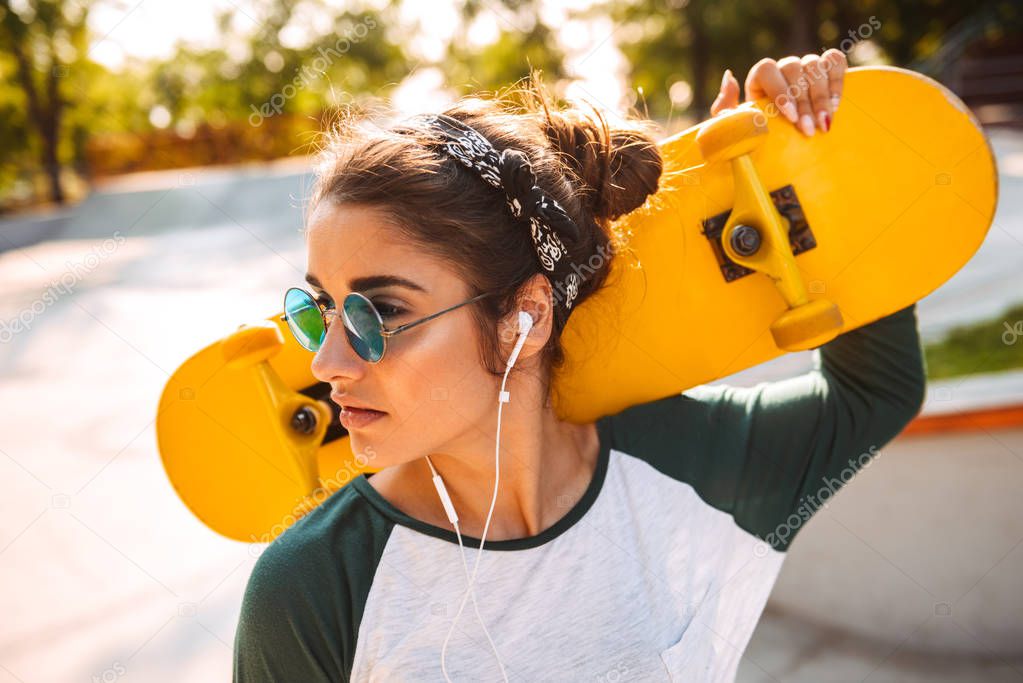 Photo of young beautiful woman walking outdoors with skateboard listening music with earphones.