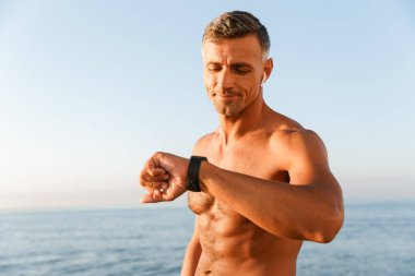 Plajda akıllı saatini kontrol kulaklık ile gülümseyen yakışıklı gömleksiz sporcu yakın çekim