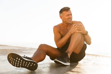 Young shirtless sportsman suffering from a knee pain at the beach clipart