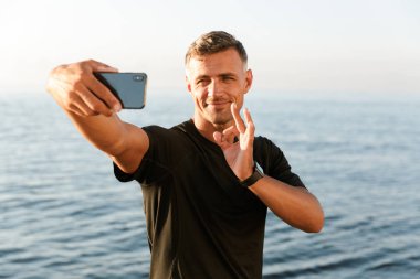 Gülümseyen yakışıklı gömleksiz sporcu sahilde duran ve ok gösterilen bir selfie alarak