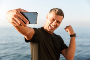 Gülümseyen yakışıklı gömleksiz sporcu sahilde duran ve pazı esneme sırasında bir selfie alarak