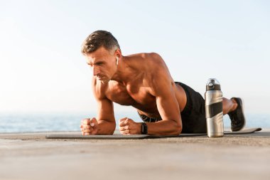 Shortless sporcu bir fitness mat plajda tahta egzersizleri yapıyor kulaklık yoğunlaştı