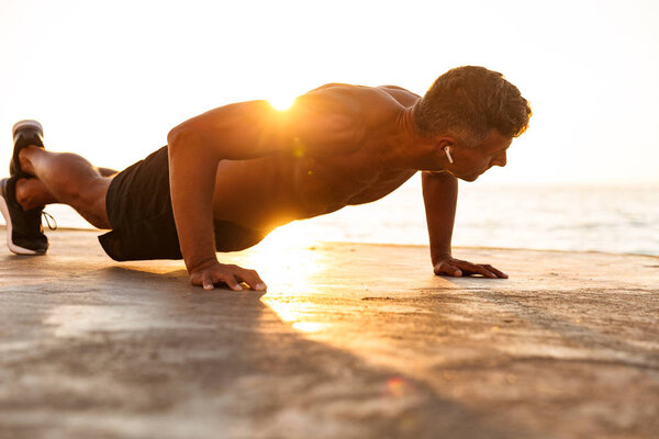 Confident sportsman with earphones doing push-ups at the beach in sunlight