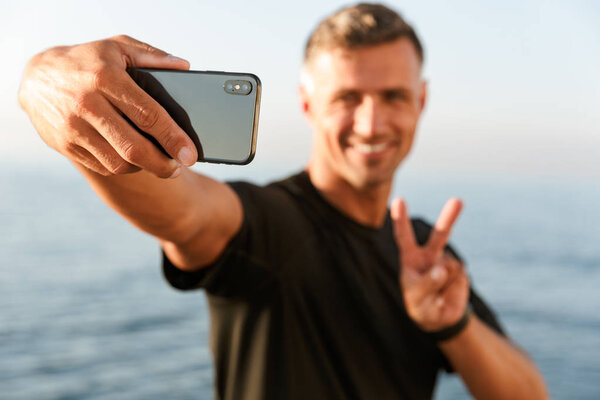 Smiling handsome shirtless sportsman taking a selfie while standing at the beach and showing peace gesture