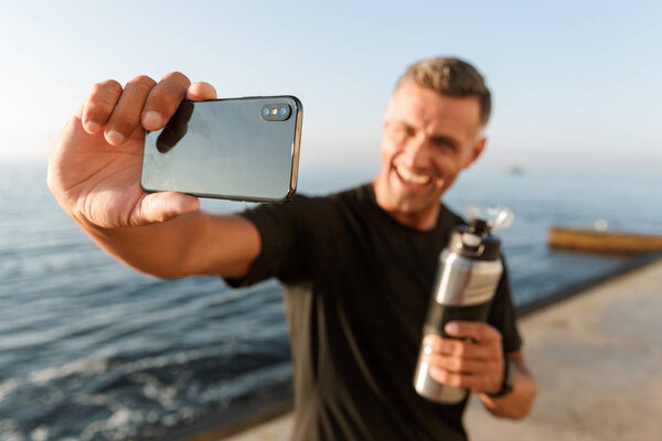 Image of handsome mature sportsman take a selfie with bottle of water on the beach.