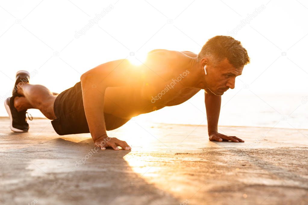 Confident sportsman with earphones doing push-ups at the beach in sunlight