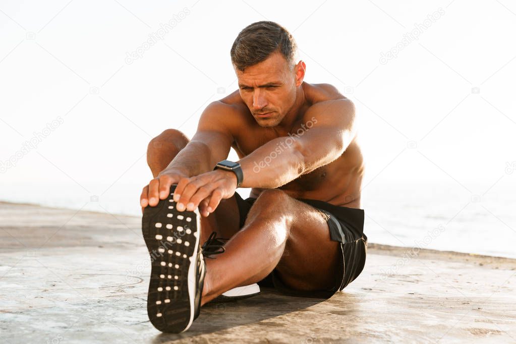 Handsome shirtless sportsman doing stretching exercises at the beach