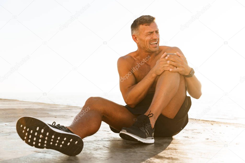 Young shirtless sportsman suffering from a knee pain at the beach