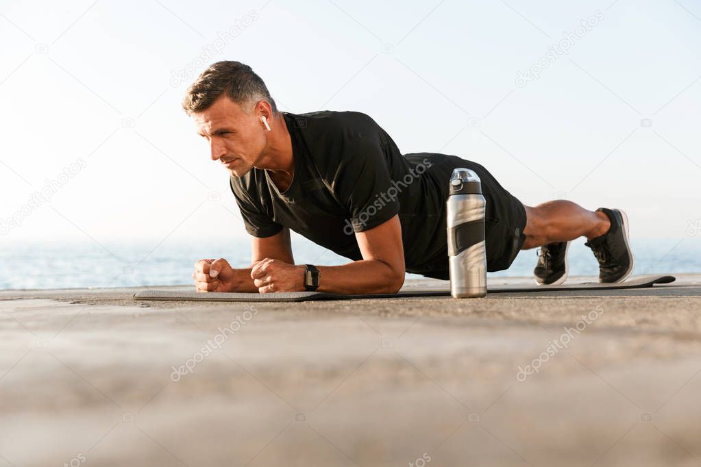 Confident sportsman in earphones doing plank exercises on a fitness mat at the beach