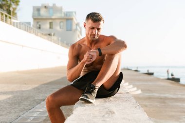 Plajda otururken akıllı saatini kontrol kulaklık ile gülümseyen yakışıklı gömleksiz sporcu